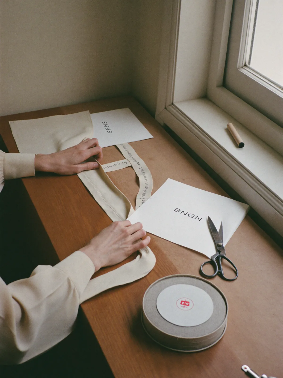 Hands pinning washed linen on the atelier cutting table, tape measure and grandmother's scissors beside
